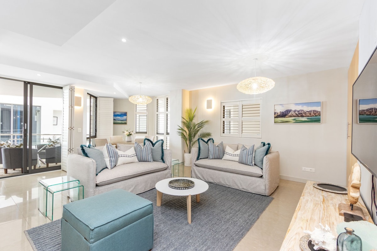 The lounge area features soft, neutral-toned sofas adorned with decorative pillows. A glass coffee table is positioned in the center, while a plush area rug adds warmth. Natural light enters through large windows, complemented by contemporary light fixtures and indoor greenery.
