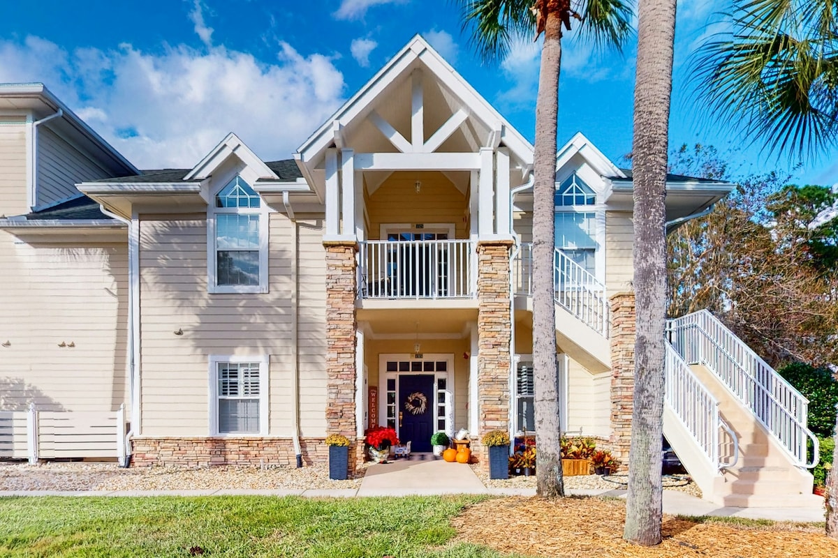 The exterior of a two-story condo is shown, featuring a prominent entrance with stone accents and a welcoming porch adorned with potted seasonal plants. Stairs lead to the second floor, framed by palm trees under a clear blue sky.