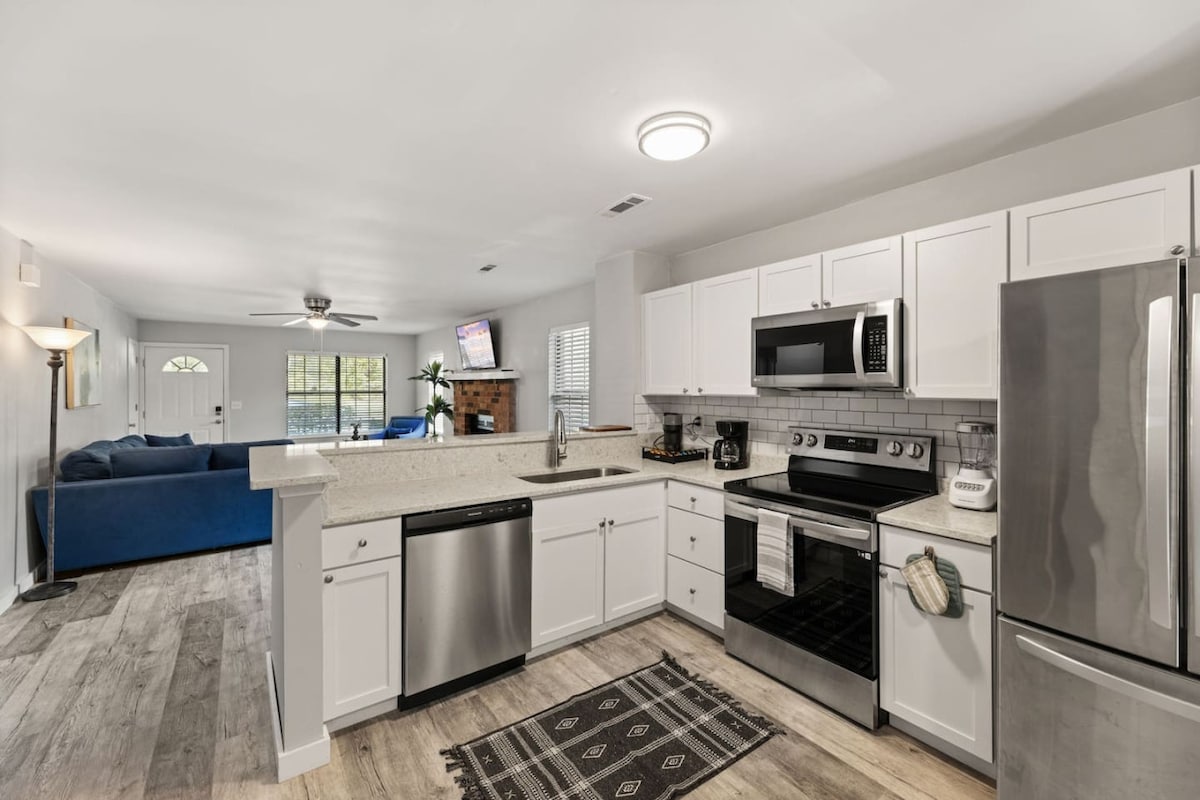 A modern kitchen features white cabinetry and stainless-steel appliances, including a dishwasher and microwave. A countertop with bar seating is visible, along with a patterned area rug. The open layout allows a view into the living area, where a blue sofa is positioned.