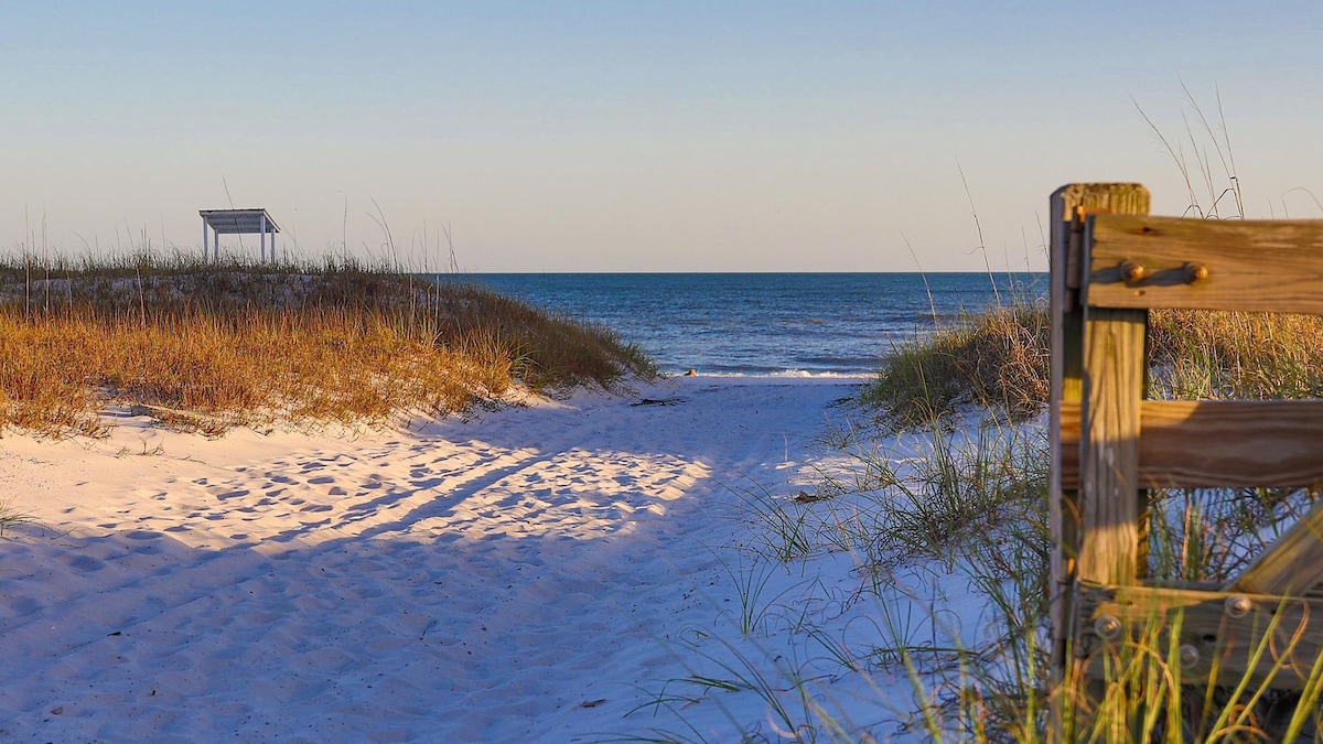 A tranquil pathway leads to the beach, bordered by gentle sand dunes and tall grass. A wooden railing frames the scene, guiding the eye toward the calm blue waters and clear sky. A gazebo structure is visible in the distance, providing a sheltering viewpoint.