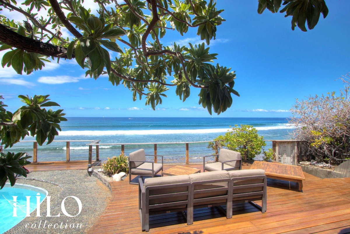 A wooden deck is seen overlooking the ocean, featuring a seating area with multiple chairs and a table. Lush greenery frames the area, and a pool is visible nearby. Clear skies and gentle waves enhance the tranquil coastal setting.