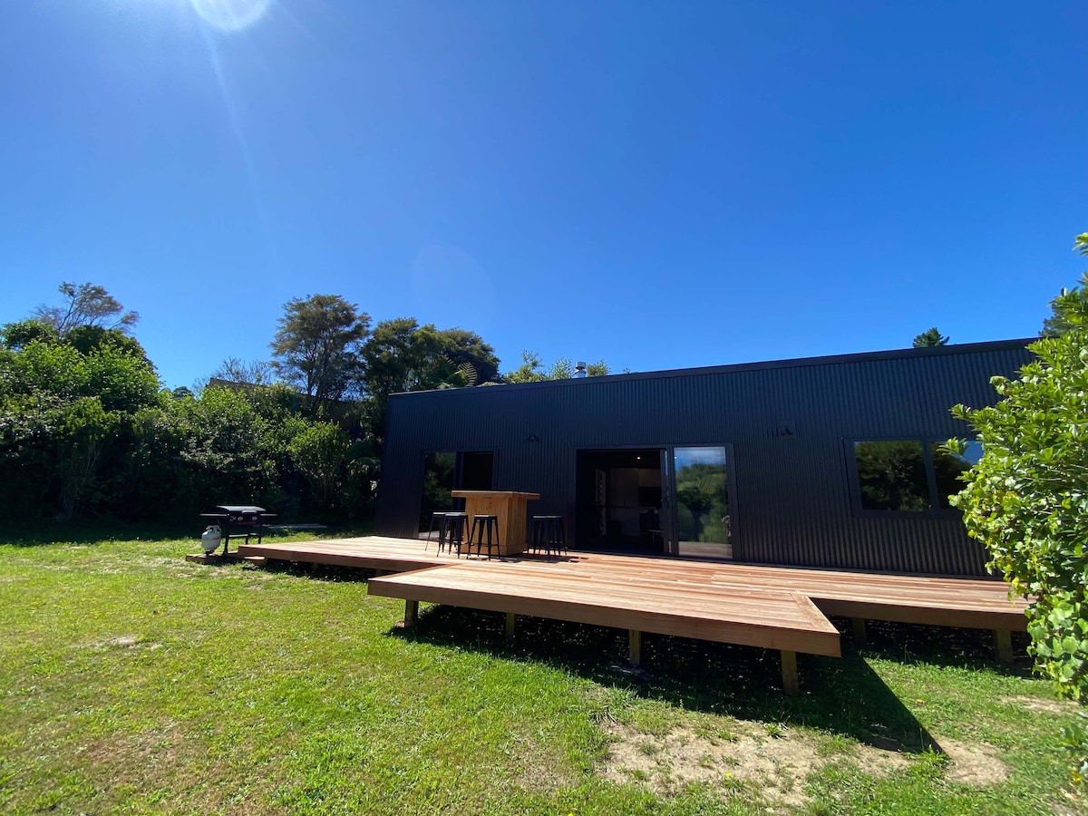 A modern holiday home is showcased against a clear blue sky, featuring a spacious wooden deck. Outdoor seating is provided at a tall table, surrounded by grassy areas and lush greenery, creating a serene outdoor space for guests to enjoy.