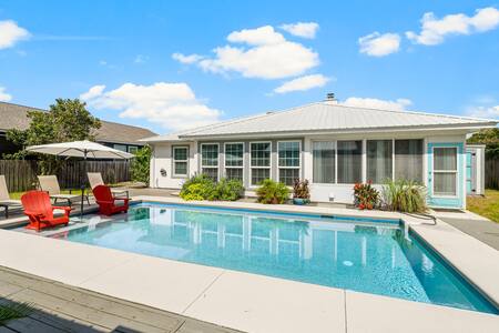 A private backyard features a heated salt-water pool surrounded by landscaped areas. Two red lounge chairs and an umbrella provide shaded seating nearby. Large windows of the house offer views of the sunny outdoor space, enhancing the inviting atmosphere of relaxation.