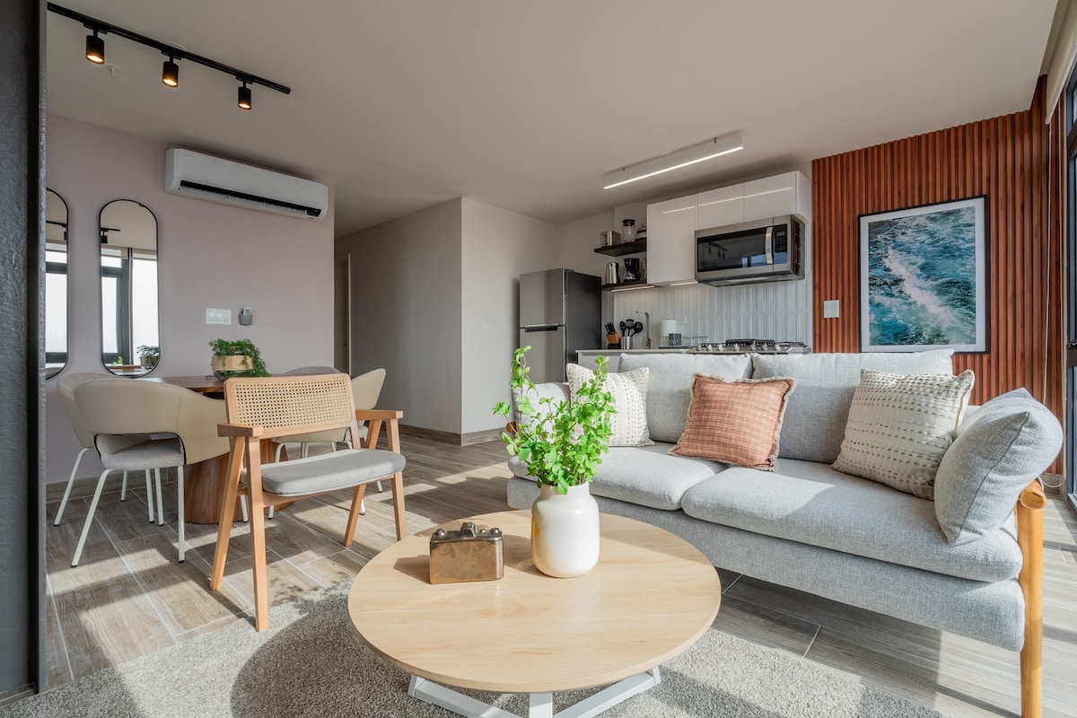 A modern living area features a light-colored sofa adorned with decorative pillows, placed across from a round wooden coffee table. Dining chairs surround a sleek table in the background, while a fully equipped kitchen with stylish cabinetry is visible to the side. Natural light streams in through the floor-to-ceiling windows.