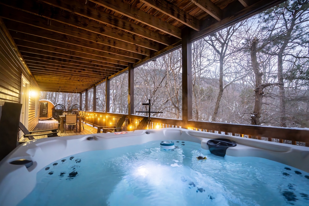 A spacious outdoor deck features a 7-person hot tub surrounded by natural wood beams. Soft glowing lights enhance the ambiance, while inviting views of the wooded surroundings are visible through large, open spaces. Comfortable seating areas add to the relaxation of this retreat.