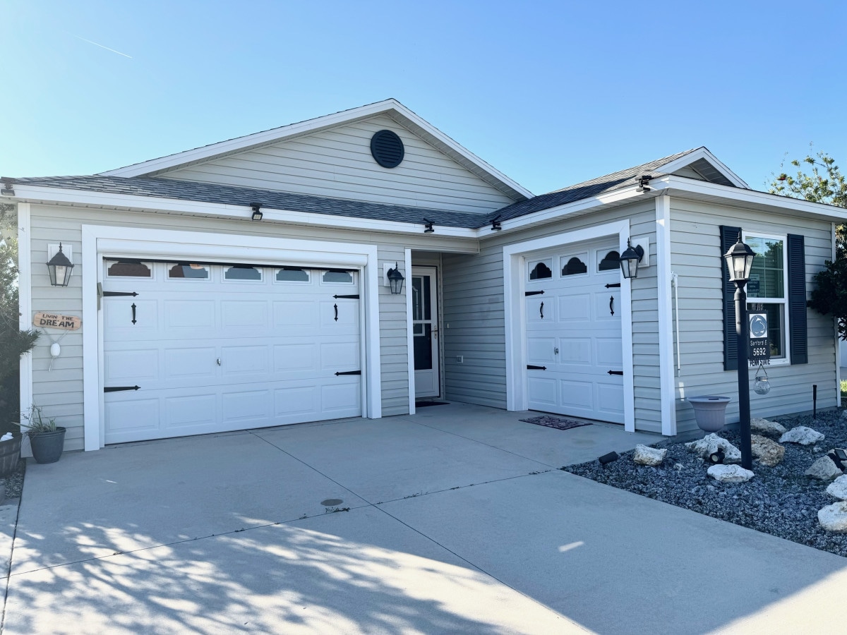 The villa's exterior showcases a contemporary single-story design with a well-maintained façade. Two white garage doors are visible, framed by neatly spaced landscaping. A welcoming front porch is complemented by fixtures that enhance the overall curb appeal under clear blue skies.