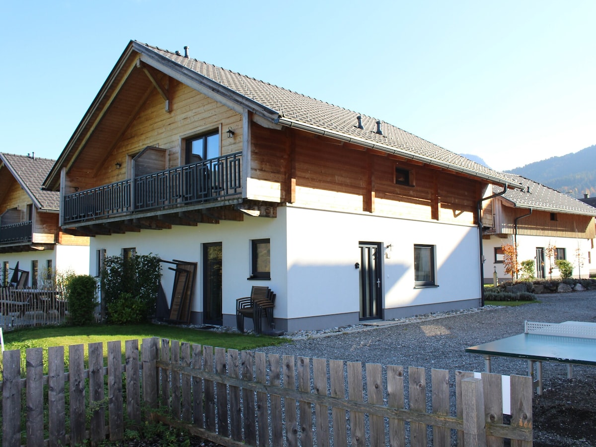 The exterior of the holiday home features a combination of wooden and white stucco finishes. A balcony with a wooden railing is visible on the upper floor, while seating and storage can be seen on the ground level. A well-maintained grassy area surrounds the structure.