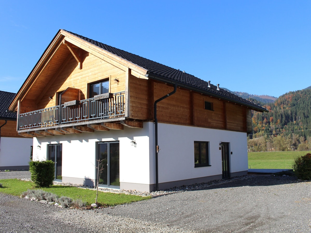 A two-story holiday home with wooden accents is set against a clear blue sky. The structure features a balcony with railing, large windows, and a landscaped area with gravel and low shrubs, offering a peaceful view of the surrounding nature.