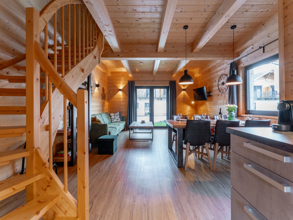 The interior space is characterized by wooden paneling and a warm ambiance. A spiral staircase leads to the upper level. A dining area features a large table set for six. A comfortable seating area is visible, complemented by large windows allowing natural light.