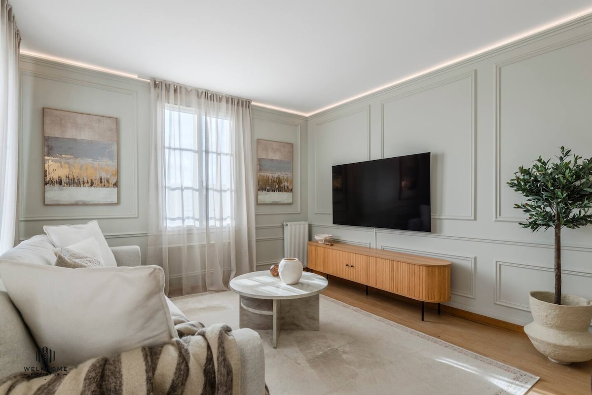A modern living area features a light-colored sofa accompanied by a decorative throw. A round coffee table sits in the center, while a flat-screen TV is mounted on the wall opposite. Natural light filters through sheer curtains, highlighting the wall art and minimalist decor.