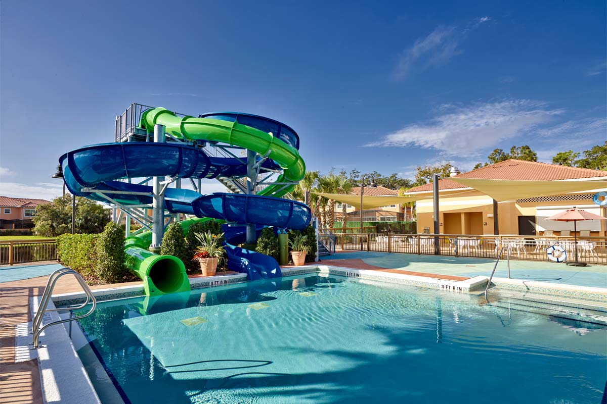 A vibrant pool area features a large blue and green waterslide curving down into a sparkling pool. Surrounding the pool, lush greenery and colorful potted plants add a natural touch. Sun loungers and umbrellas are visible along the deck, inviting relaxation and enjoyment.
