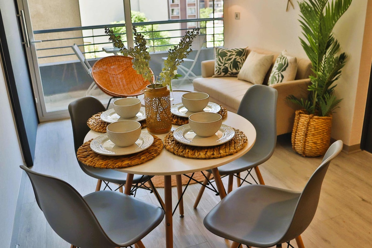 A modern dining area features a round table set for four, complete with bowls and textured placemats. Four stylish chairs surround the table, while a comfortable sofa is visible in the background. Decorative plants and a woven basket add a touch of greenery.