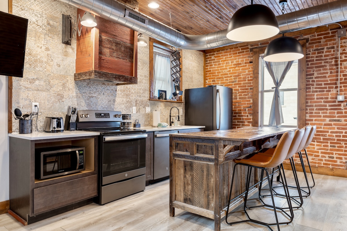 The kitchen area features exposed brick walls and wooden accents. Stainless steel appliances, including a refrigerator and oven, are visible. A wooden kitchen island with seating for four is present, complemented by stylish pendant lighting overhead. A microwave is also located on the countertop.