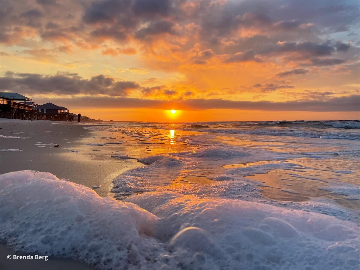 The image captures a serene beach scene at sunset, with soft waves rolling onto the shore. The sky is painted in shades of orange and pink, reflecting in the wet sand. Fluffy clouds enhance the tranquil atmosphere as the sun descends toward the horizon.