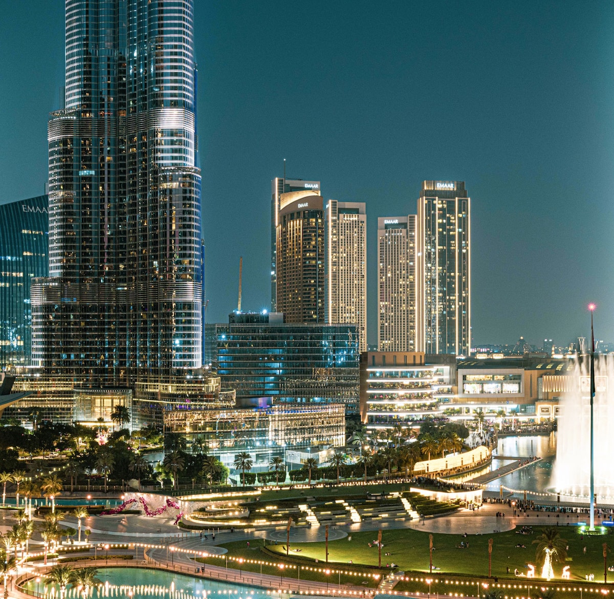 A night view of Downtown Dubai showcases the illuminated Burj Khalifa alongside modern skyscrapers. The vibrant landscape features lush greenery and an array of lights reflecting off the buildings, creating a lively urban atmosphere. The nearby water fountain display is visible, adding to the scenic charm.