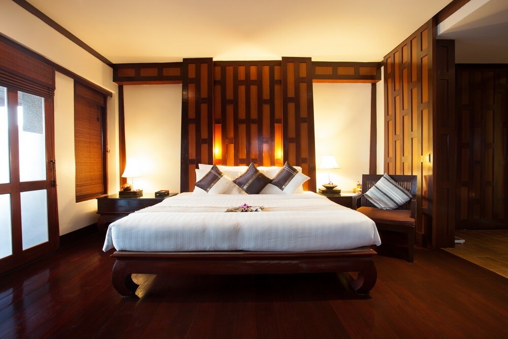 A spacious bedroom features rich wooden accents with warm lighting. A large bed is centered against a decorative wall, flanked by nightstands with lamps. Natural light enters through windows covered by bamboo shades, adding a serene touch to the ambience.