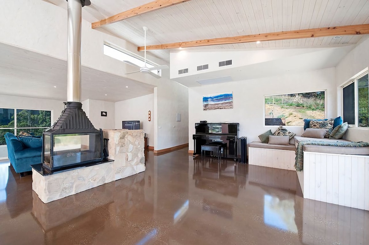 An open-plan living area is highlighted by a wood-burning fireplace and large windows that frame mountain views. Comfortable seating arrangements, including a blue couch and built-in lounge, invite relaxation. A piano adds a musical element to the space, while polished concrete flooring enhances the modern aesthetic.