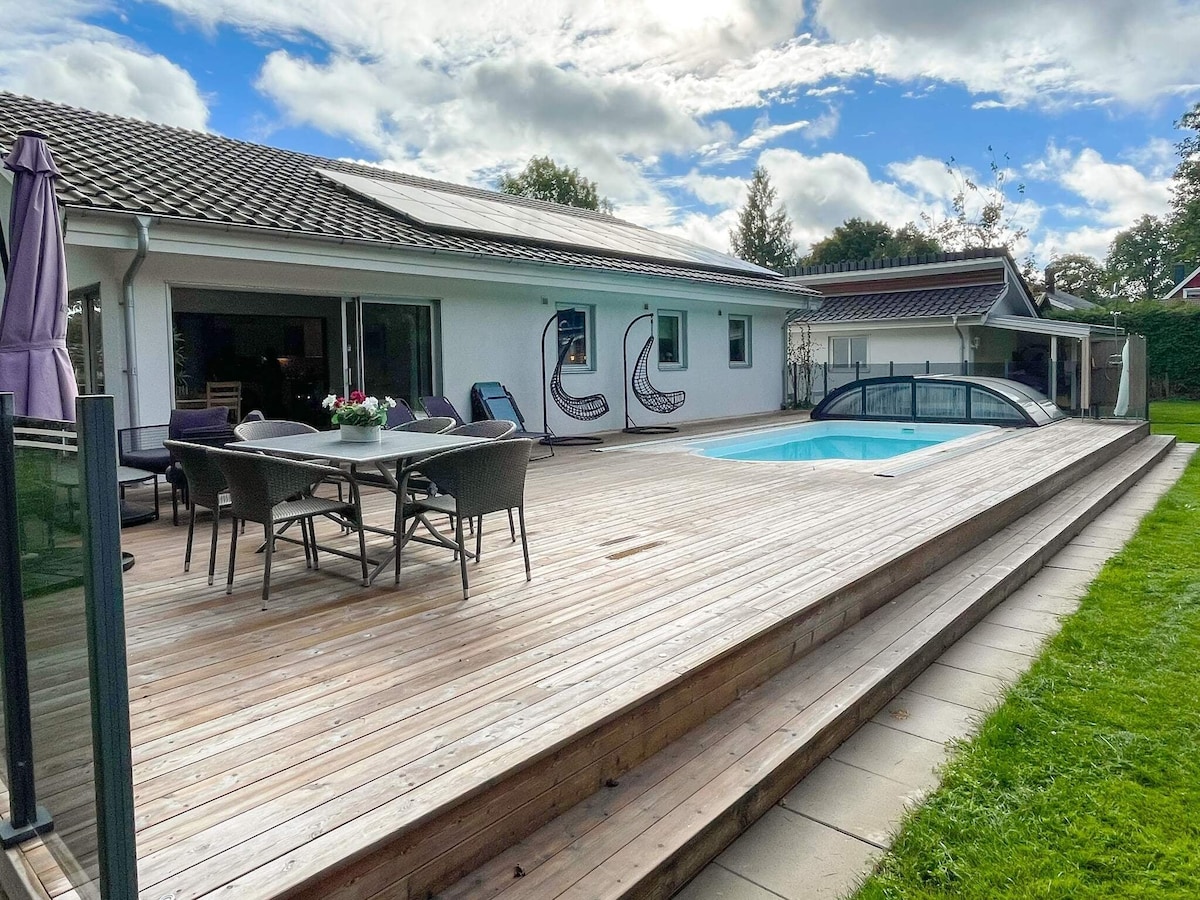 A wooden deck surrounds a small pool, offering ample space for outdoor seating. Chairs and a dining table are placed on the deck. The smooth surface transitions to a well-maintained grassy area, while large windows reveal the interior of the home.