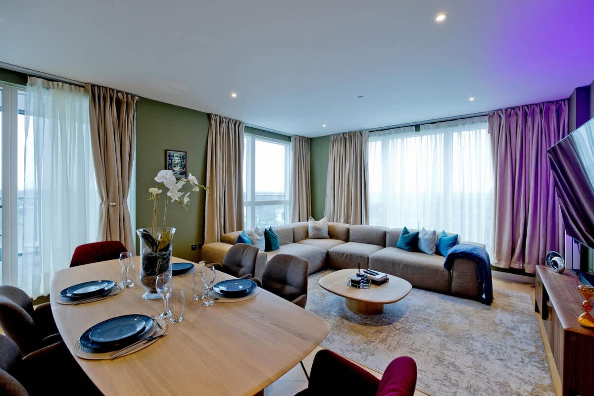 A spacious living and dining area is depicted, featuring a large sectional sofa with decorative cushions, framed by floor-to-ceiling windows that allow natural light. A wooden dining table set for six sits adjacent to the sofa, complemented by an elegant coffee table and a large TV.