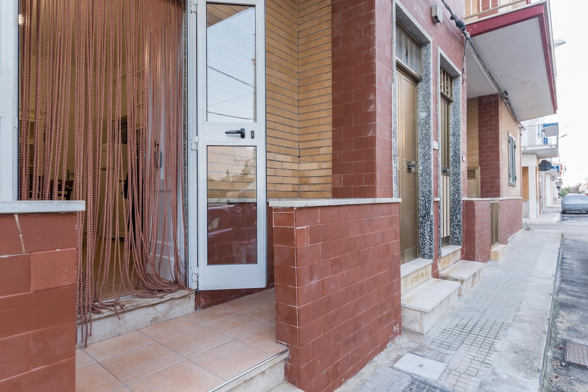 The exterior of a building features a brick facade, with a clear entrance door that opens to a welcoming space. The entrance is adorned with decorative ropes, and a small set of stairs leads to the door. Sidewalks are visible along the street.