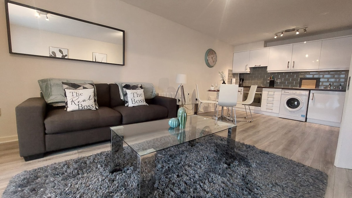 A modern living area is featured, with a comfortable dark sofa adorned with decorative pillows. A glass coffee table sits atop a plush rug, reflecting natural light from the space. The open kitchen and dining area include sleek cabinets, a dining table with chairs, and a washing machine.