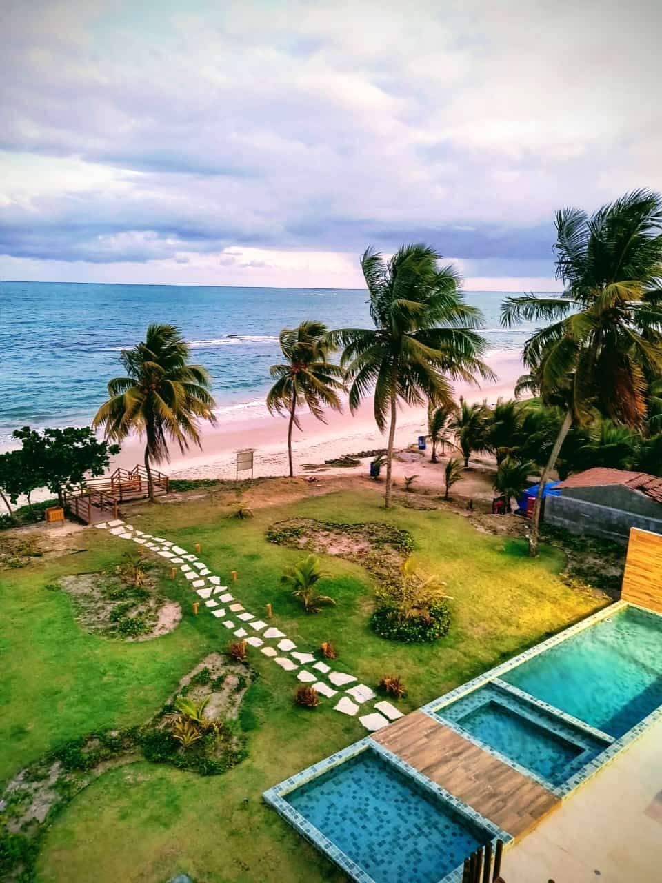A serene beachfront scene showcases lush greenery with palm trees swaying gently in the breeze. A stone path leads toward the sandy shore, while a tranquil pool glistens under the soft light, inviting relaxation. Waves can be seen lapping at the beach just beyond.