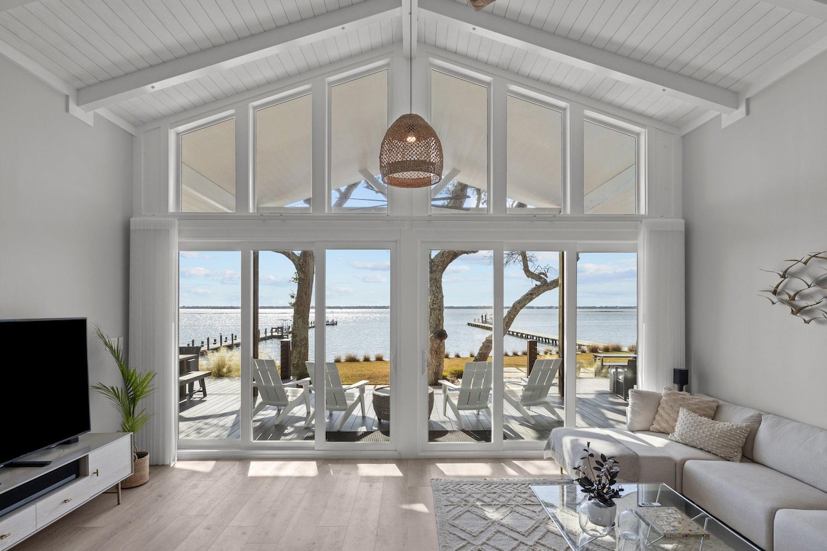 A bright living area features a high, vaulted ceiling and expansive glass doors that open to scenic waterfront views. Comfortable seating is arranged around a glass coffee table, with a decorative plant adding a touch of greenery. Natural light floods the space, creating an airy atmosphere.