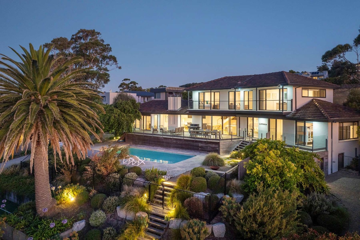 A spacious coastal villa is showcased at dusk, with a well-lit pool and outdoor seating area. Lush gardens and a palm tree frame the scene, while large windows reflect the tranquil surroundings, inviting relaxation in a serene setting.