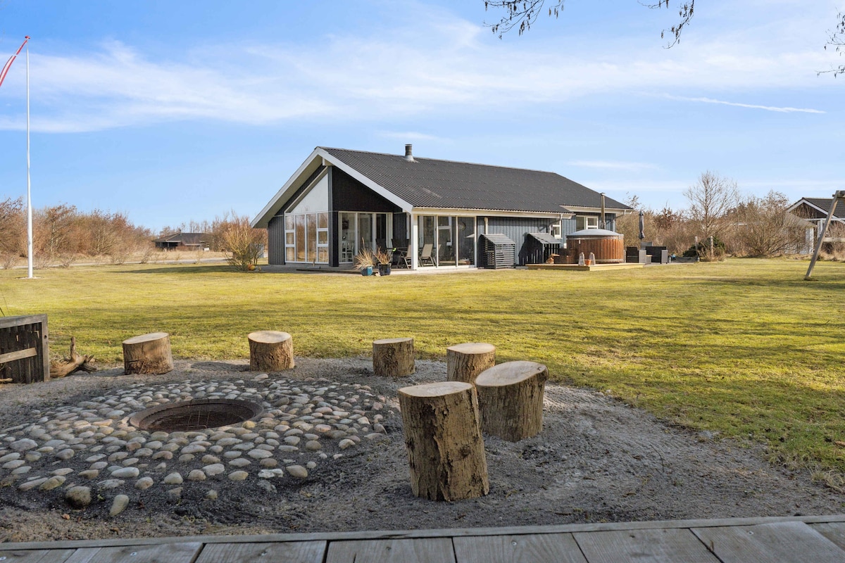 A spacious backyard is surrounded by natural landscaping, featuring a fire pit made of stones and wooden seating. The holiday home stands in the background, showcasing large glass doors and a sleek roof design. A hot tub is visible nearby, adding to the outdoor amenities.