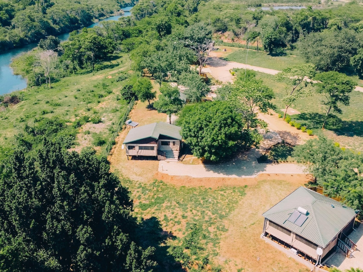 An aerial view captures the chalet's outdoor surroundings, featuring landscaped greenery and nearby river. Two structures are visible, surrounded by a peaceful natural setting, with pathways connecting them. Trees dot the landscape, enhancing the serene environment.