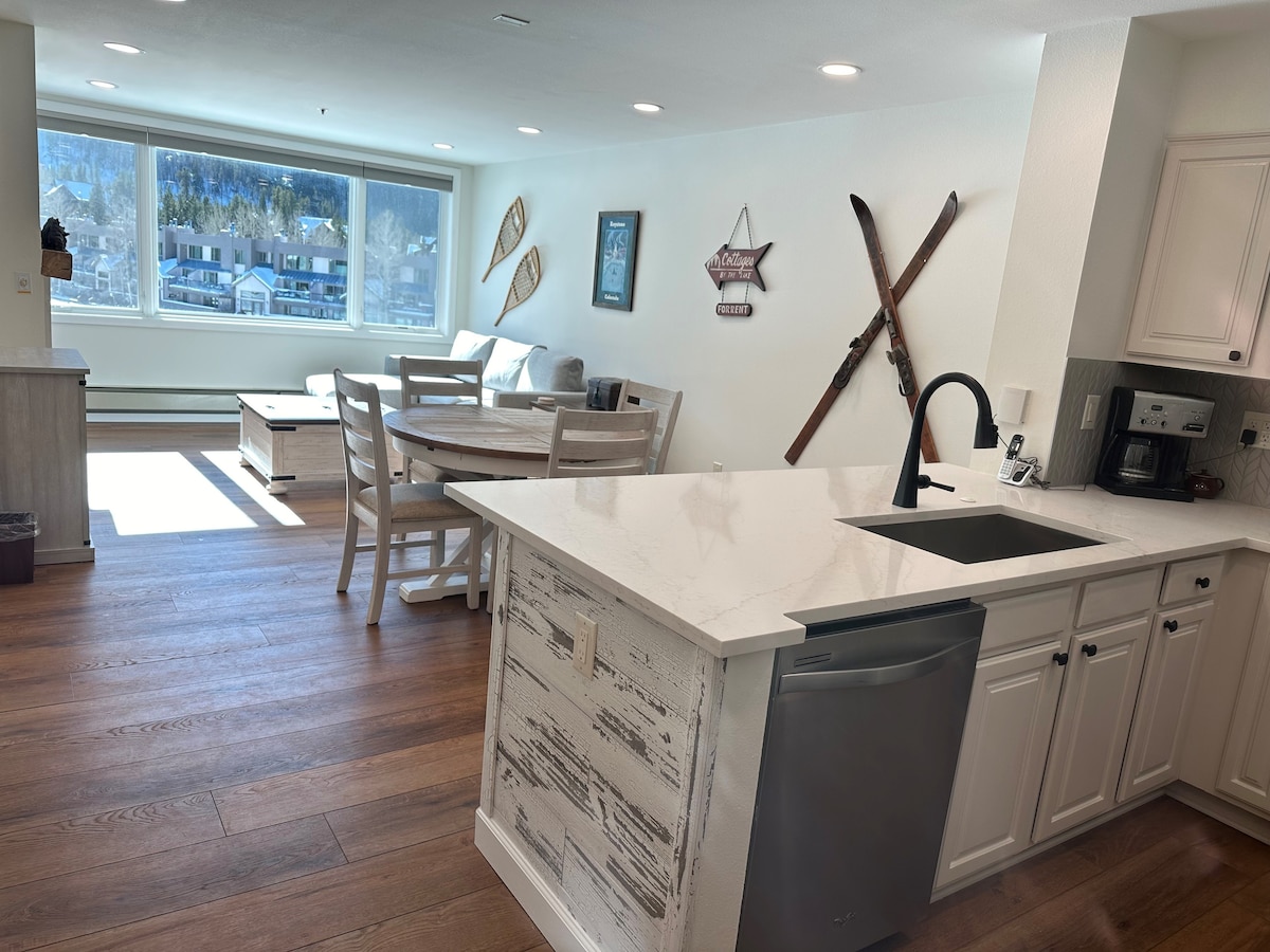 An open kitchen area features modern appliances and a spacious island with seating. Natural light streams in from large windows, illuminating a cozy dining space with a round table. Wall decor includes vintage skis and art, enhancing the alpine ambiance.