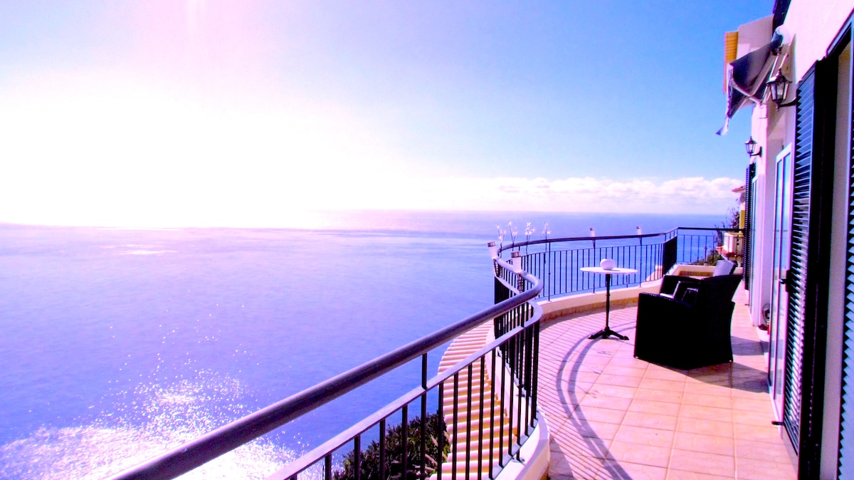 The private balcony offers sweeping 180-degree views of the ocean, with tiled flooring and a curved railing enhancing the open atmosphere. A small table and two chairs provide a cozy spot for outdoor dining or relaxation in the warm sunlight.