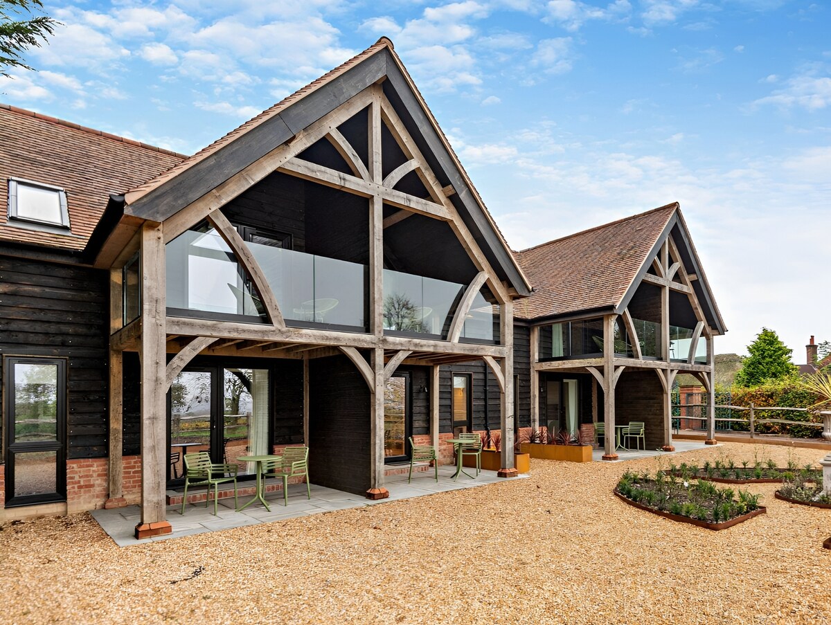 A modern barn conversion is showcased, featuring a spacious exterior with large glass panels and wooden supports. Outdoor seating is arranged on a stone patio, surrounded by a landscaped garden, creating an inviting space that blends seamlessly with the natural surroundings.