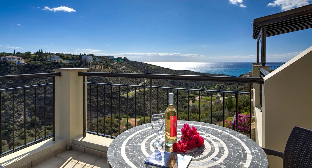 A scenic terrace overlooks the ravine and sea, featuring a round table arranged with a bottle of white wine, two glasses, and a bouquet of vibrant flowers. The stunning backdrop includes rolling hills and the sparkling sea under a clear blue sky.