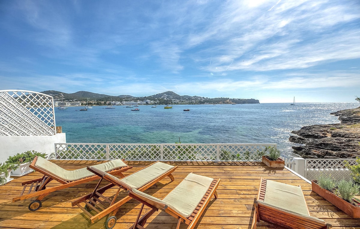 An open terrace features four sun loungers arranged on warm wooden decking, overlooking the sparkling sea. The expansive view captures boats gently bobbing on the water, framed by a vibrant blue sky with wispy clouds.