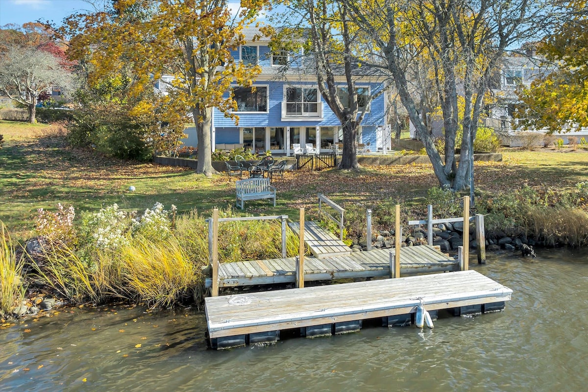A private dock extends into the calm water, featuring a spacious platform perfect for boat access. The background showcases a large, multi-level home painted in soft blue, surrounded by autumnal trees displaying vibrant yellows and oranges, adding warmth to the scenic waterfront setting.