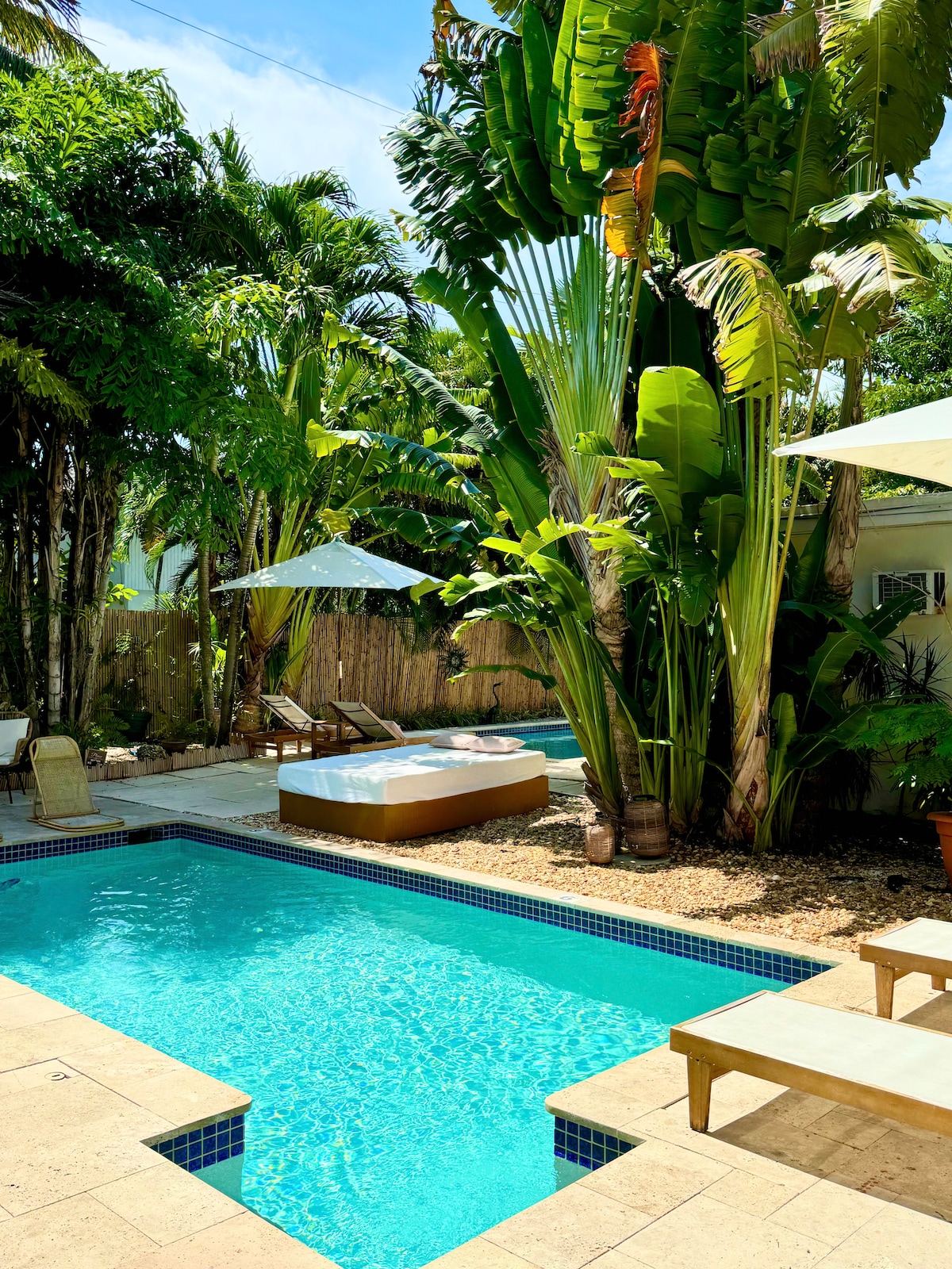 A serene outdoor space showcases two inviting pools amidst lush tropical foliage. Comfortable lounge chairs are positioned nearby, and large umbrellas provide shade. The atmosphere is bright and inviting, complemented by the natural beauty of vibrant green plants.