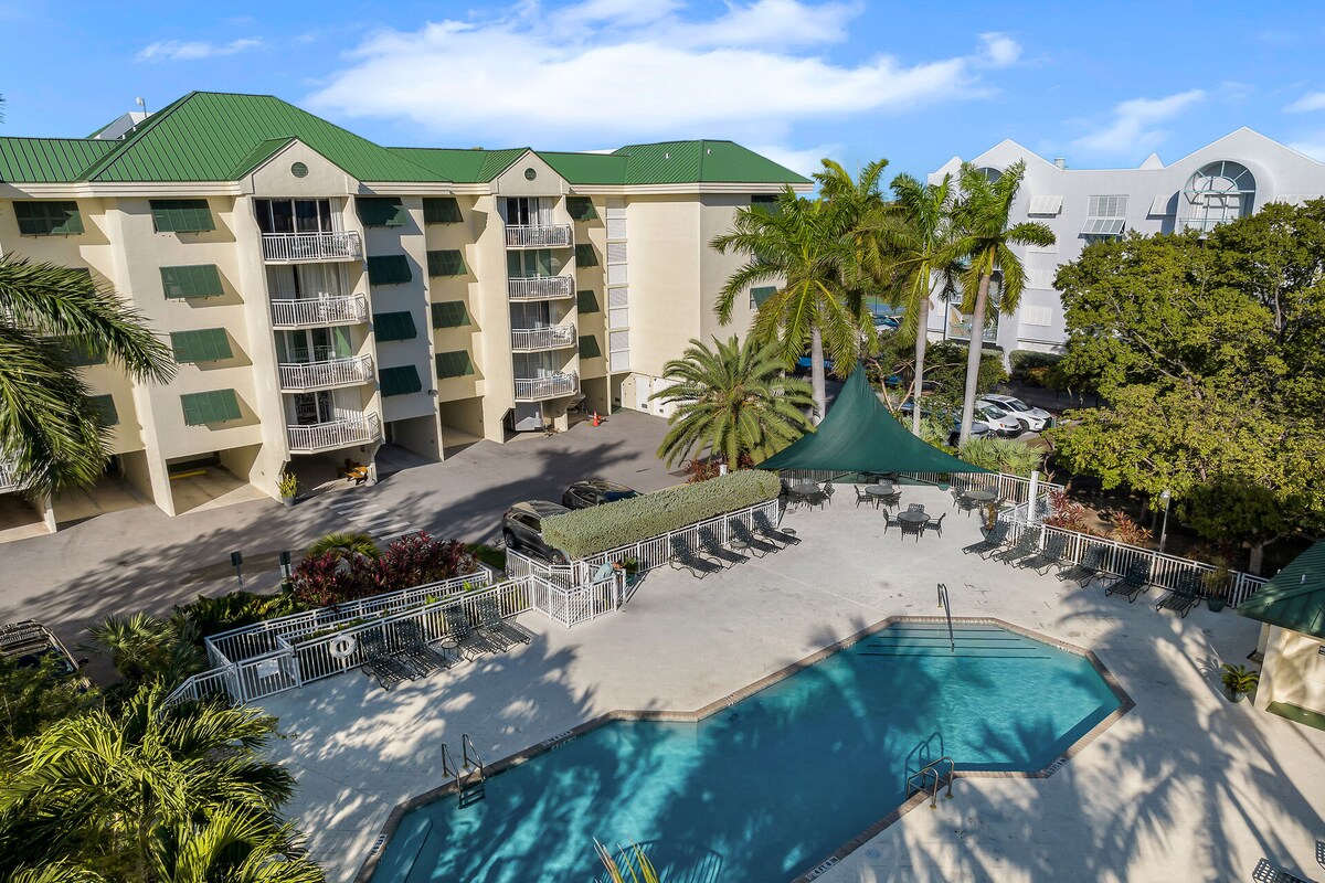 The image showcases a multi-story building with a green roof, surrounded by tropical palm trees. A shared pool is visible in the foreground, accompanied by lounge chairs and a shaded area. Parking spaces are indicated in the background, providing convenient access.