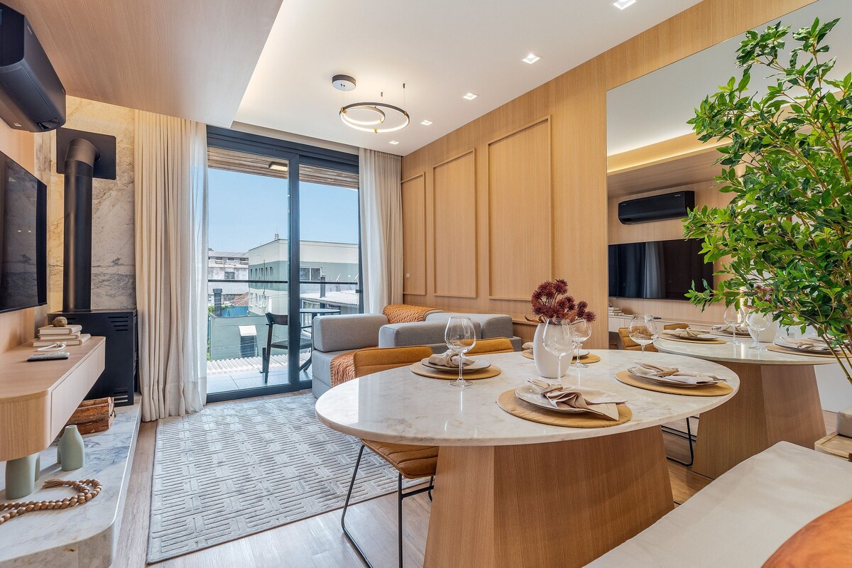 The living area features a modern design with a large window opening to a view. A neutral-toned sofa is positioned next to a television and a fireplace. A circular dining table, set with tableware, complements the warm wood accents and greenery throughout the space.