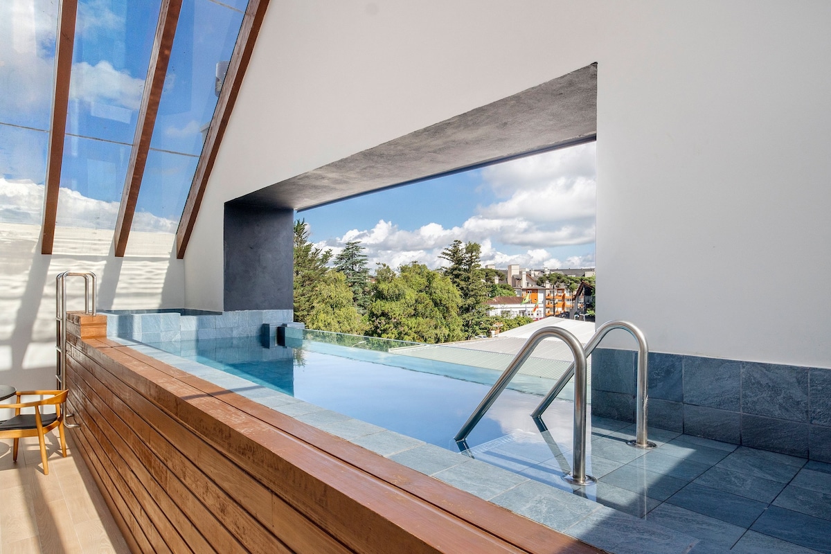 An indoor pool is showcased with a panoramic window offering views of the surrounding landscape. Natural light illuminates the space, highlighting wooden accents and tiled surfaces. Pool steps provide access, while the clean lines and modern design create a serene environment.