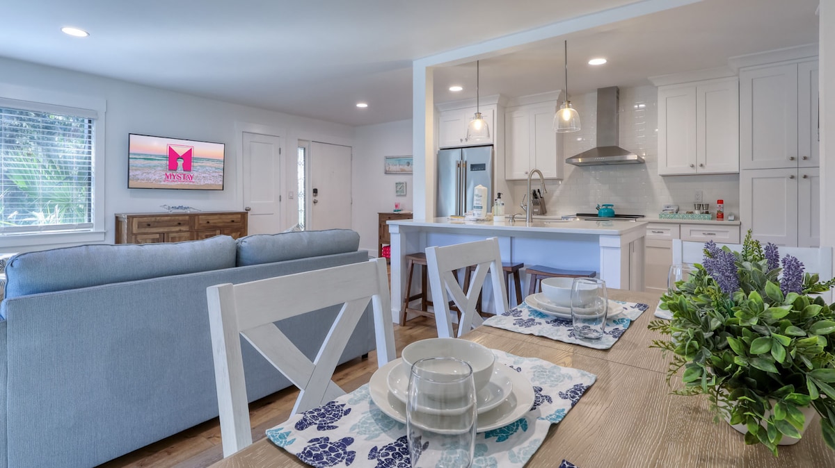 A light-filled open-concept living space features a comfortable sofa facing a mounted Smart TV. A dining table is set with dishware, and a kitchen with modern stainless appliances and quartz countertops is visible in the background. Soft, coastal colors create an inviting atmosphere.