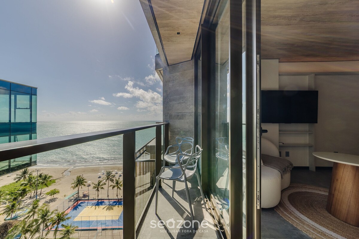 A balcony offers a side view of the sea, with sunlight reflecting off the water. Two outdoor chairs are positioned for relaxation, while a spacious living area is visible through the glass doors, creating a bright and open ambiance.