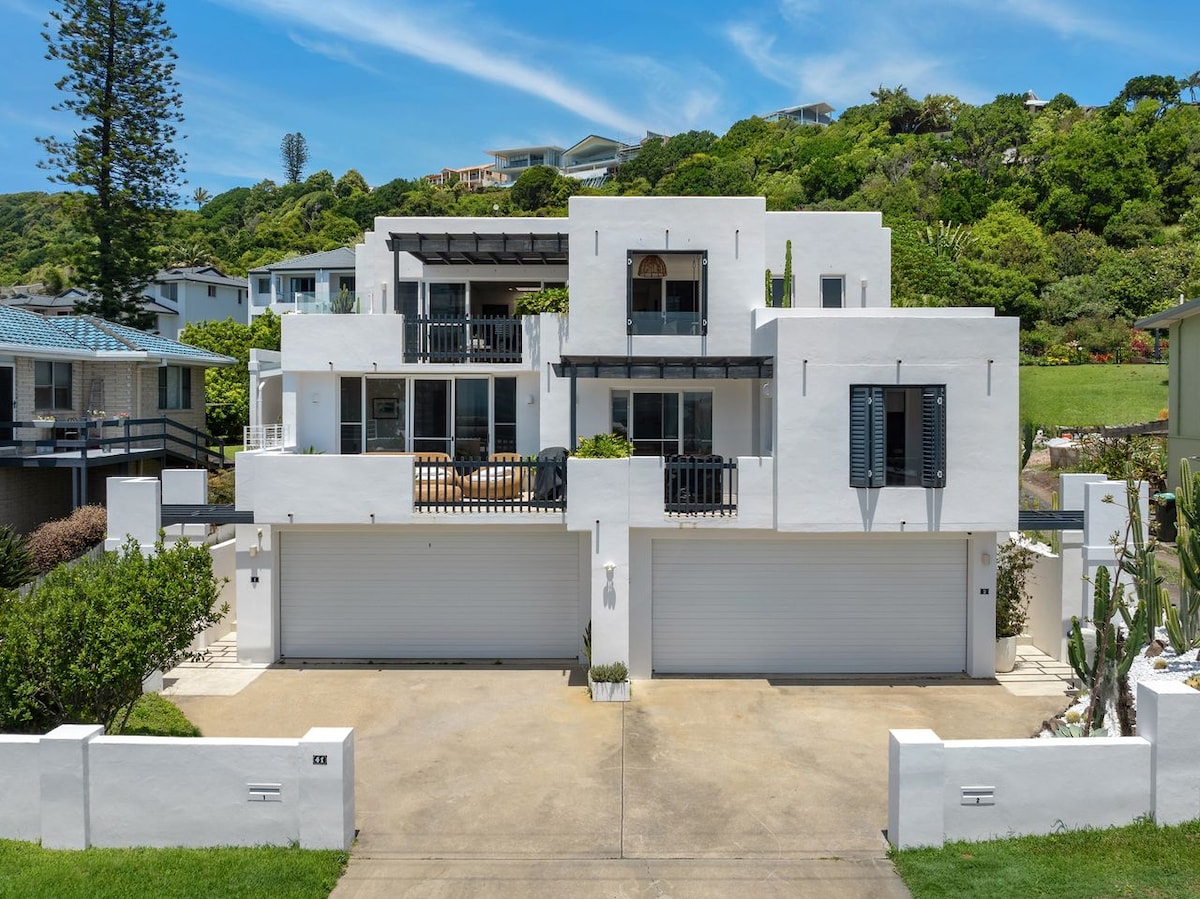 The Mediterranean villa presents a clean white facade, accented by dark shutters. A spacious balcony extends from the upper level, offering comfortable seating. Two large garage doors are visible at the base, surrounded by landscaped greenery and native plants, complementing the coastal setting.