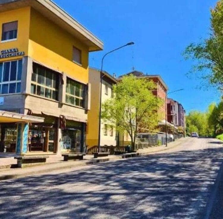 A view of a street lined with colorful buildings, showcasing a yellow structure with large windows. Trees with fresh green foliage provide a natural contrast to the building colors, and a gently sloping road leads into the scene.