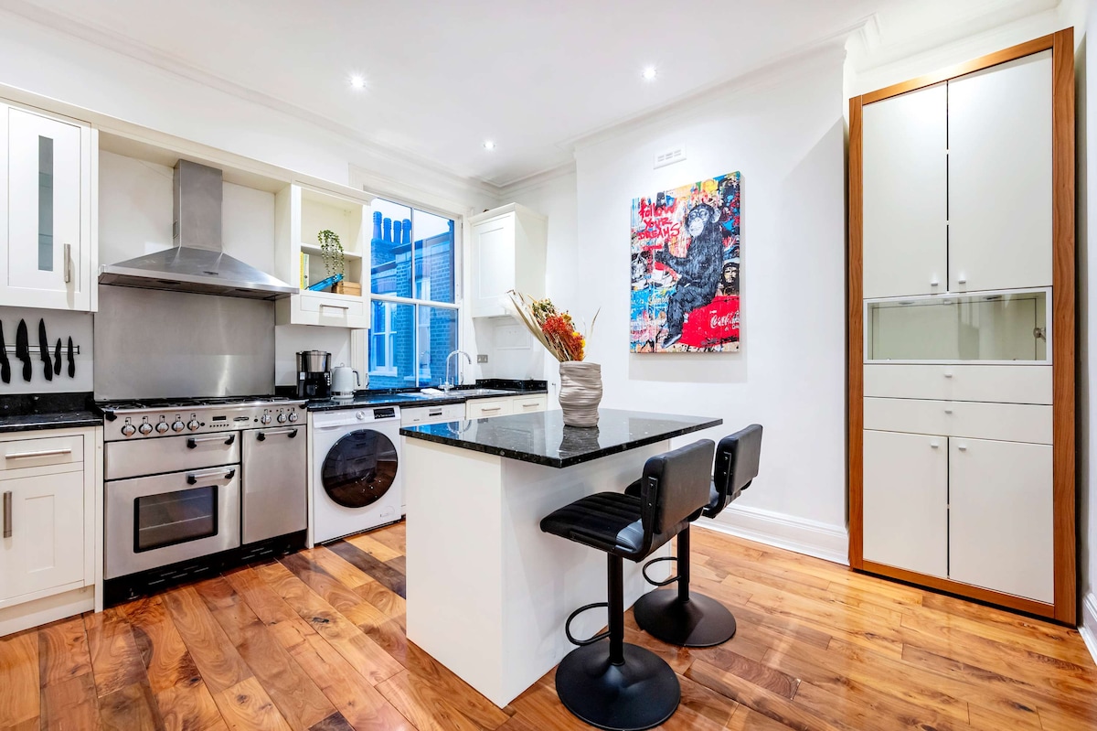 The kitchen features modern appliances including a stainless steel stove and washer, presented alongside a striking piece of artwork on the wall. The island includes two stools, and natural light streams in from the window, illuminating the wooden floors and sleek cabinetry.