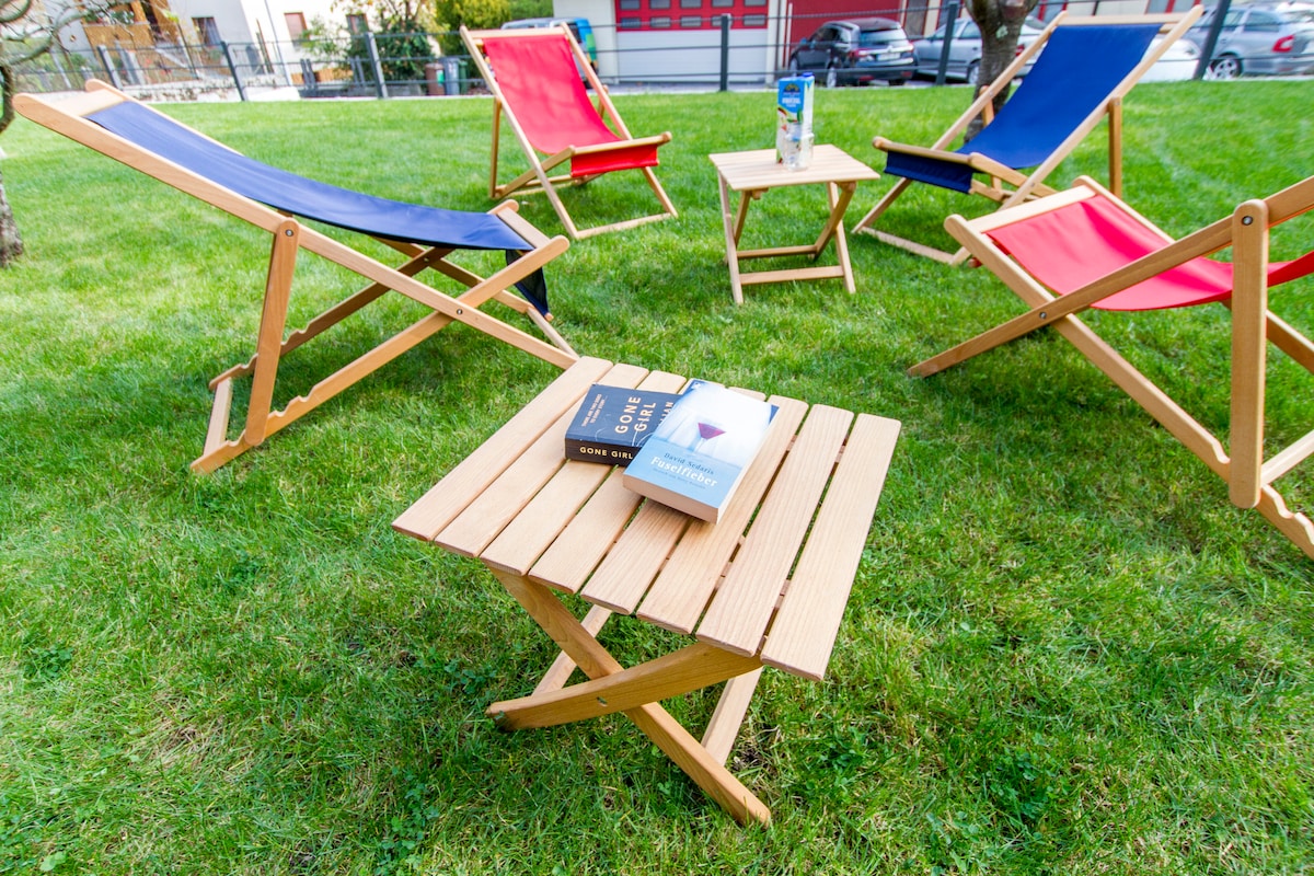 A spacious garden area is furnished with wooden deckchairs, including red and blue options, arranged around a small wooden table. Two books rest atop the table, set on vibrant green grass, inviting relaxation in a serene outdoor environment.