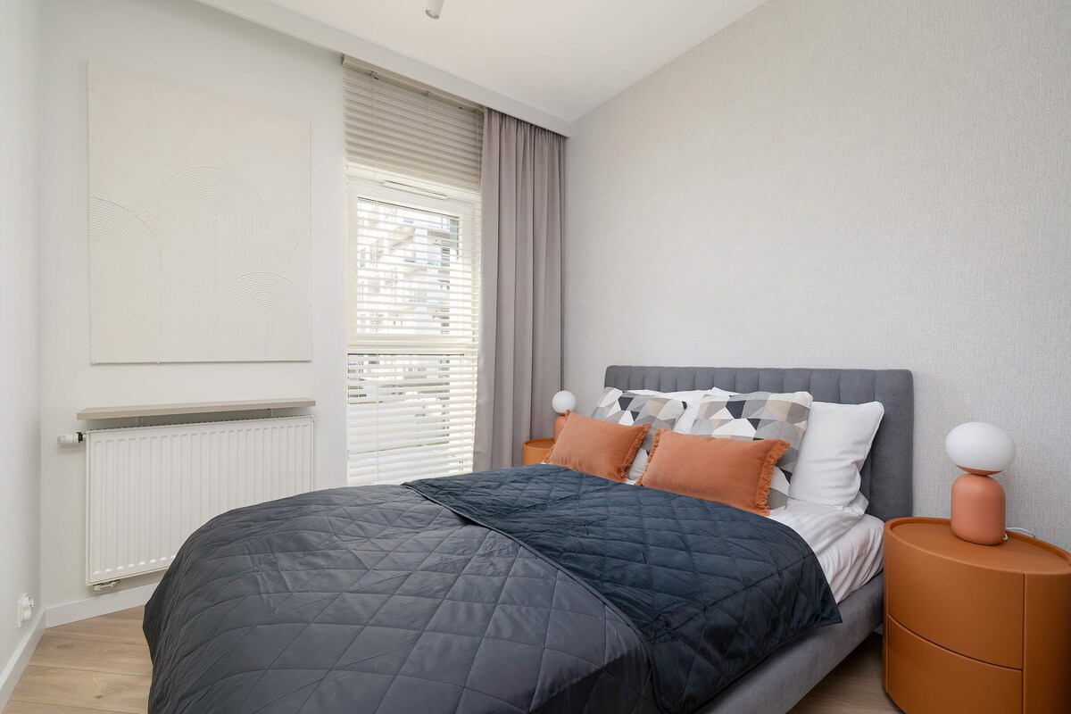 Მყუდრო საძინებელი კომფორტული ნაცრისფერი ორადგილიანი საწოლით, რომელსაც აქვს ლურჯი თეთრეული, ხის გვერდითი მაგიდა და ფართო გარდერობი.