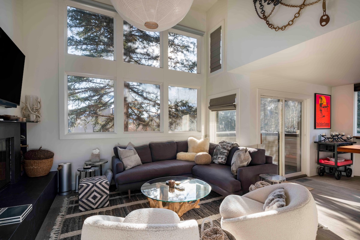 A spacious living area features large windows allowing natural light to fill the room, with views of surrounding trees. A dark sectional sofa is complemented by a coffee table, while cozy armchairs provide additional seating. A modern fireplace and access to the outdoor deck enhance the area.