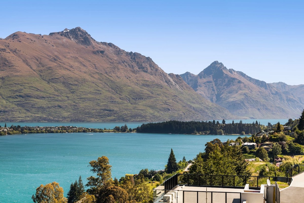 A panoramic view of a tranquil lake surrounded by majestic mountains is visible. The vibrant blue water is framed by lush greenery and the distant peaks, creating a serene natural landscape under a clear sky.