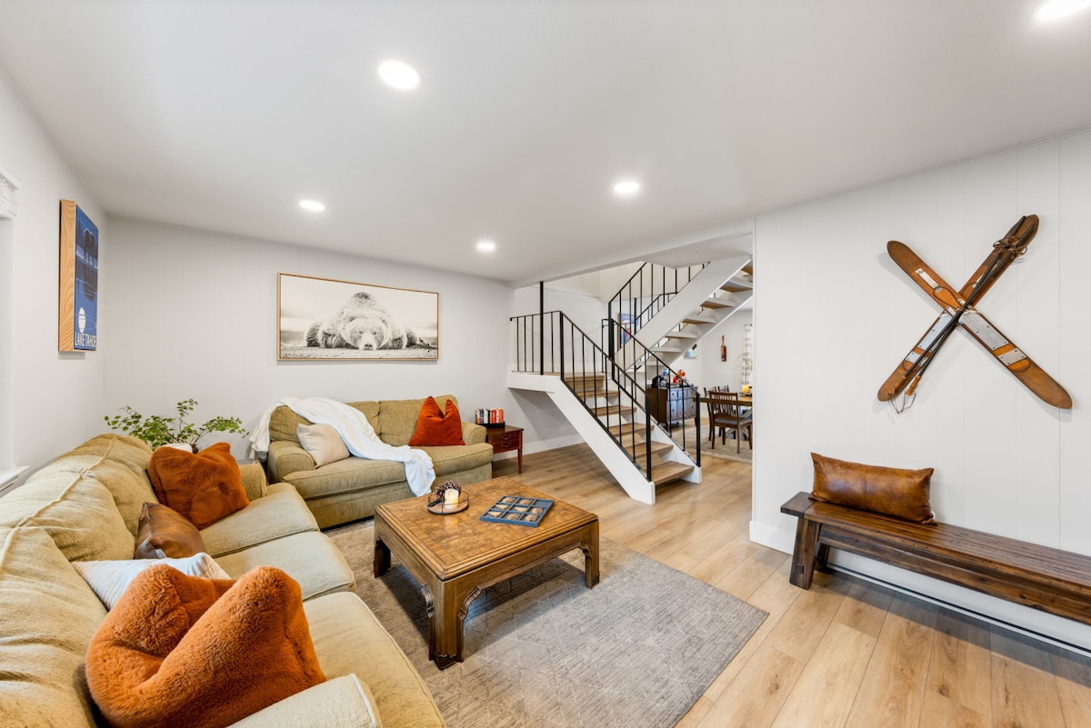 A spacious living area is highlighted by a large, L-shaped couch with soft pillows. A wooden coffee table sits in the center, complemented by decorative items. Light-colored walls and recessed lighting enhance the bright atmosphere while wooden accents and vintage skis add character.